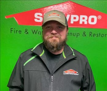 Male employee with light brown beard wearing a green hat and black jacket in front of a green wall