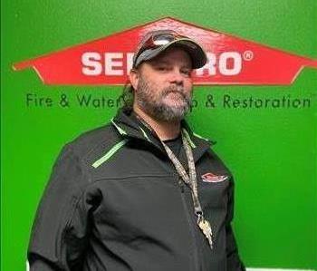 Male employee in black jacket with a hat and sunglasses and a salt and pepper grey beard in front of a green background.