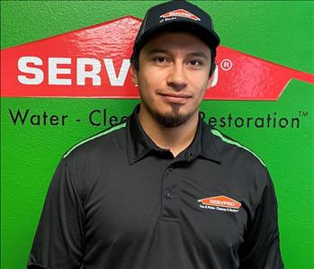 Male employee with a dark brown goatee and black hat in a black polo shirt in front of a green wall.
